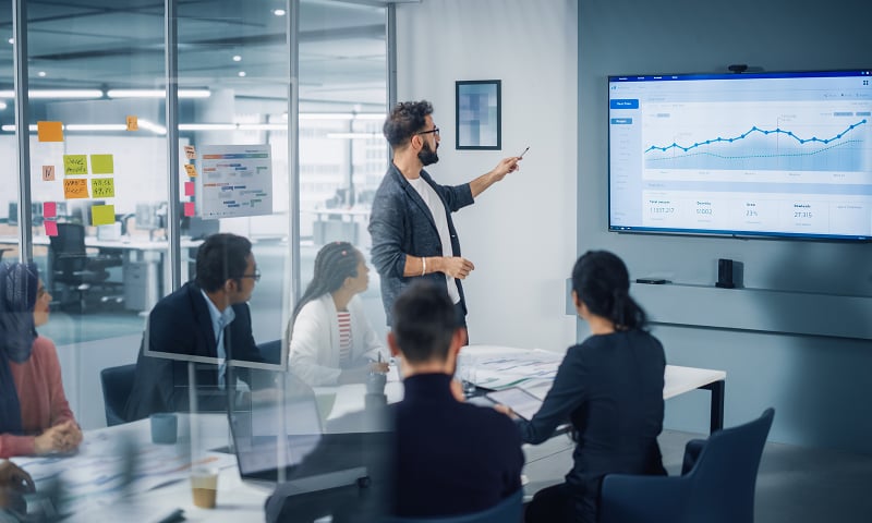 折れ線グラフが表示された大きなモニターの前でプレゼンをする男性の様子
